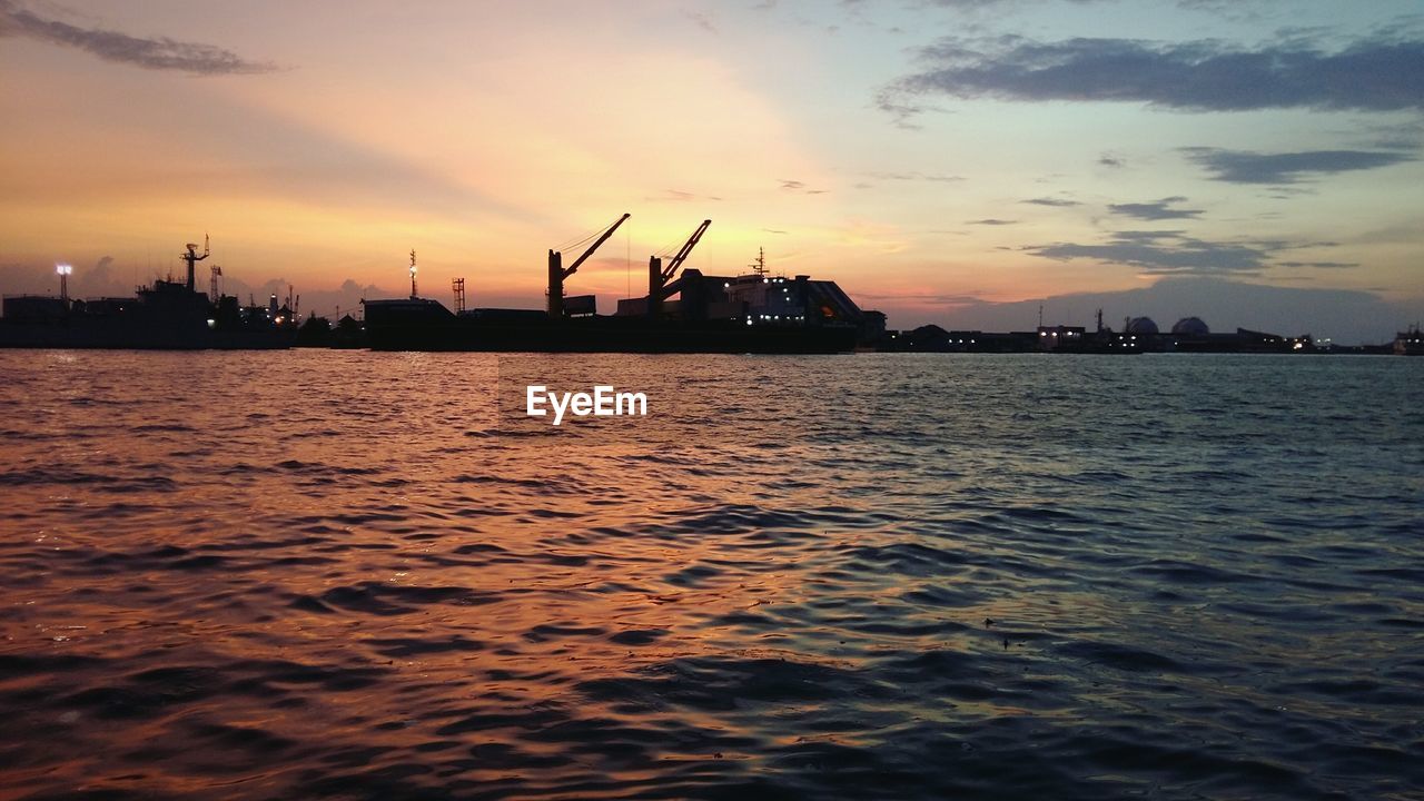 SILHOUETTE CRANES ON SEA AGAINST ORANGE SKY