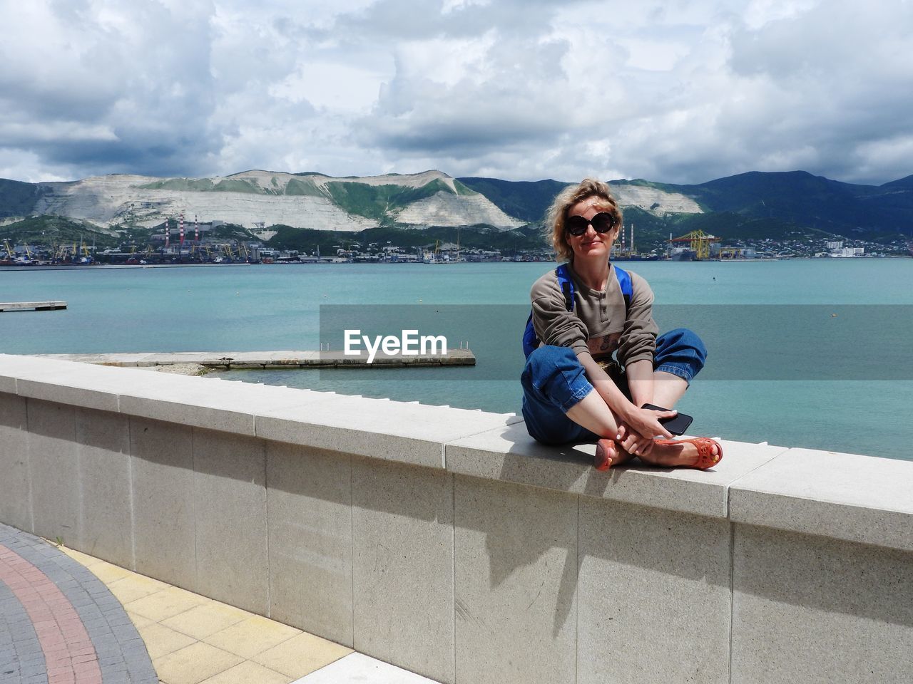 Side view of woman sitting on retaining wall by sea against sky