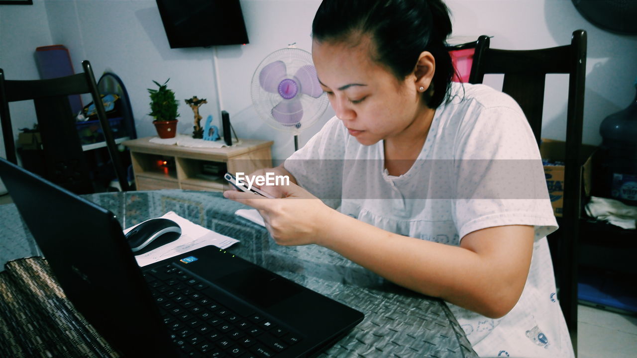 Young woman using phone in front of laptop on table at home