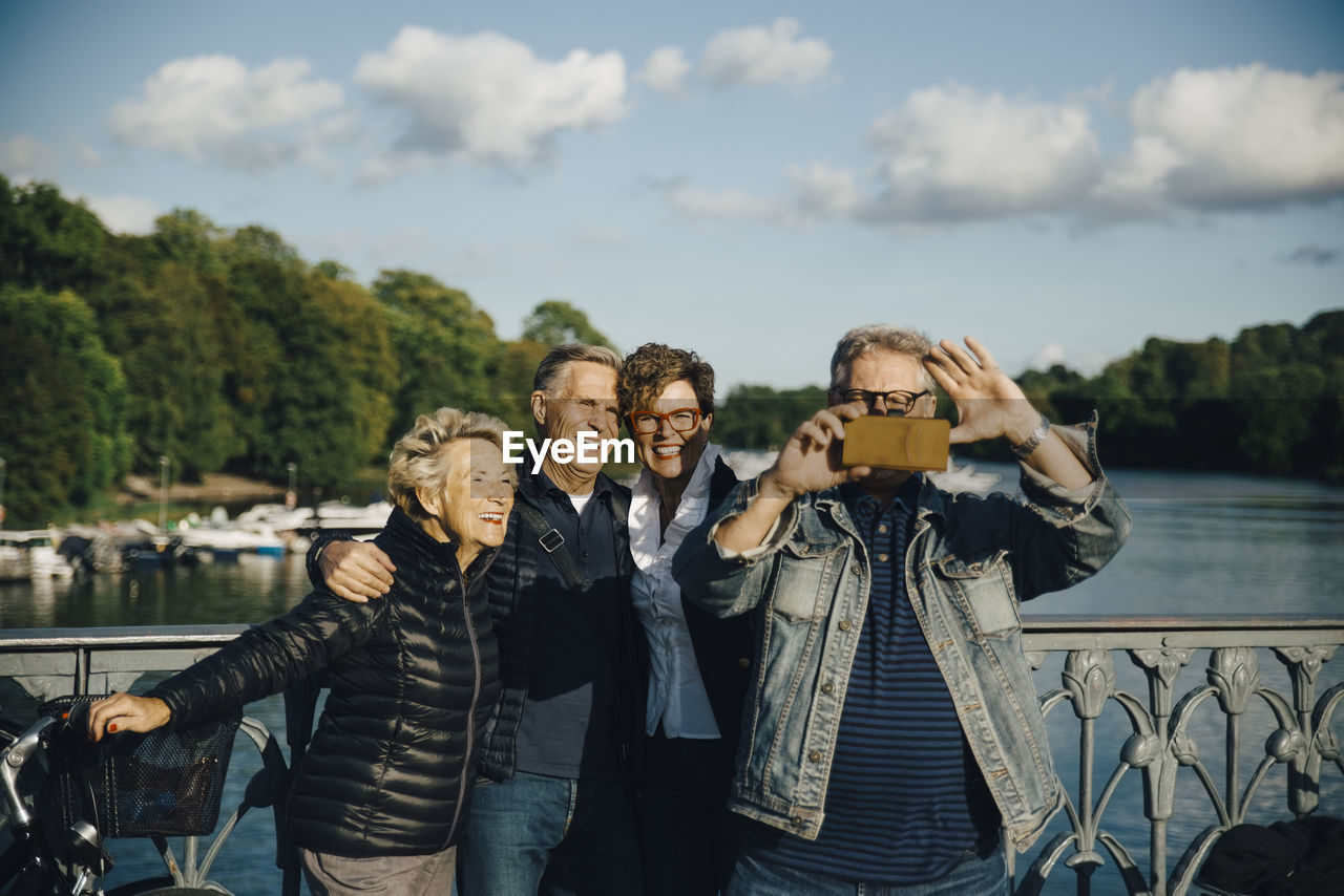 Smiling male and female friends taking selfie with smartphone by railing of bridge