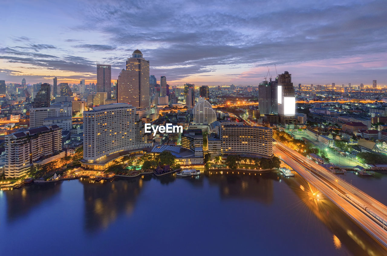 River by illuminated cityscape against sky during sunset