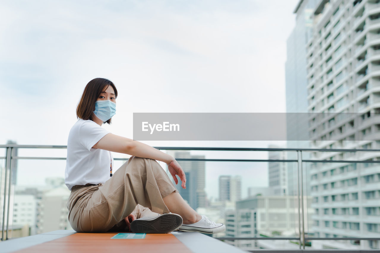 Solo asian woman wear protective mask during outdoor break and relax at rooftop with city background