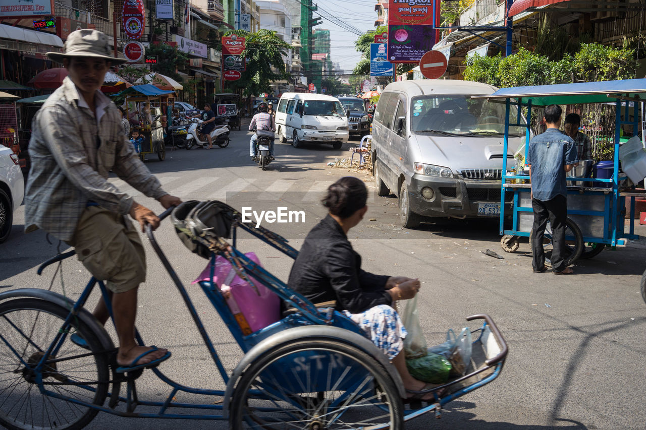 transportation, mode of transportation, city, street, rickshaw, vehicle, architecture, city life, car, group of people, bicycle, adult, men, land vehicle, motor vehicle, road, traffic, city street, transport, women, travel, mode of transport, building exterior, full length, lifestyles, day, on the move, motorcycle, outdoors, tricycle, built structure, casual clothing, driving, motion, public transportation, nature