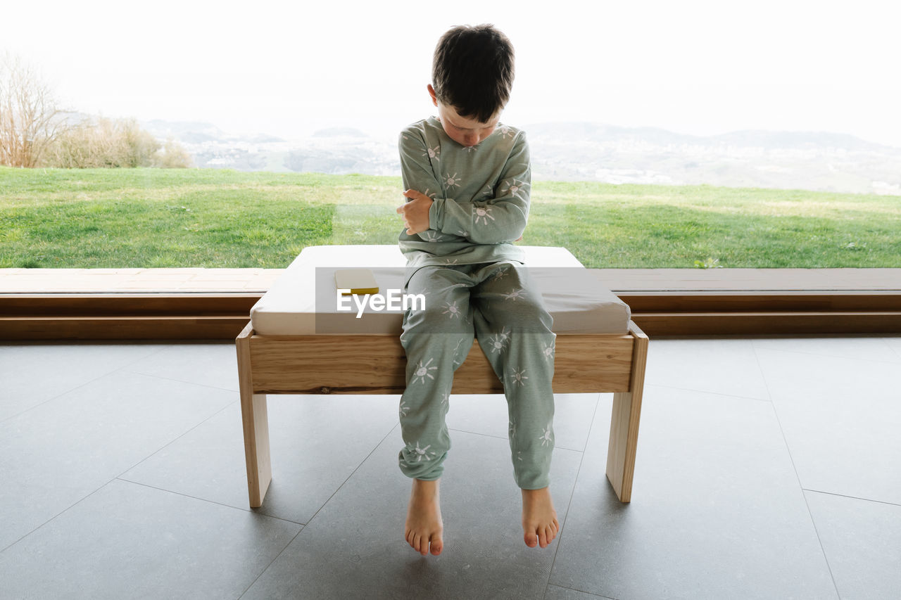 Unhappy boy in pajama sitting on pouf in light room on morning time near modern cellphone to faceless