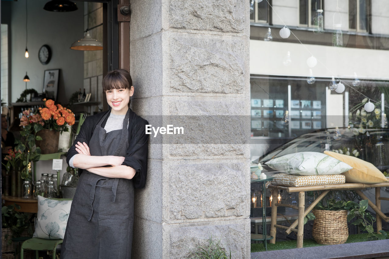 Portrait of smiling confident young female entrepreneur standing with arms crossed at store doorway