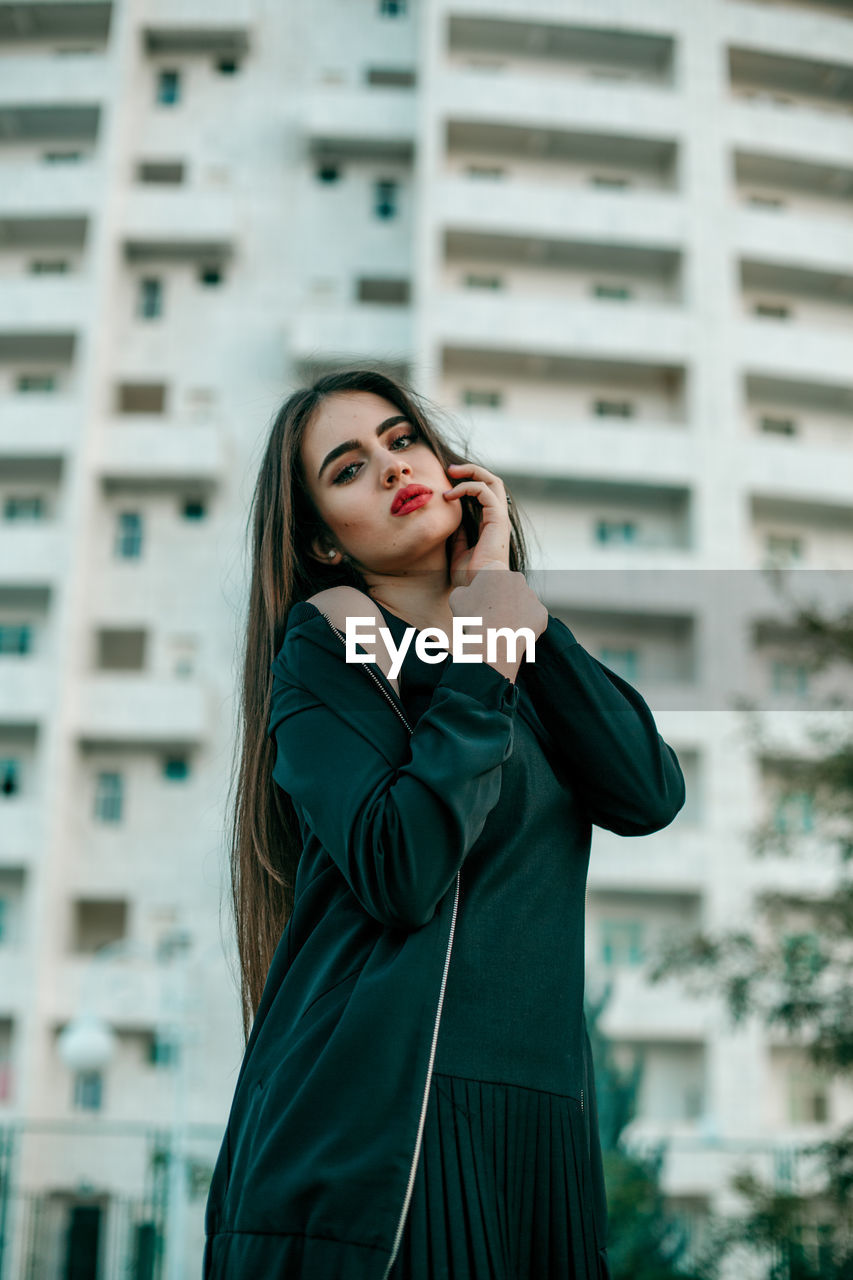 YOUNG WOMAN STANDING AGAINST BUILDING