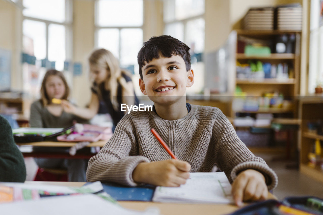 Smiling male pupil holding pencil while sitting at desk in classroom
