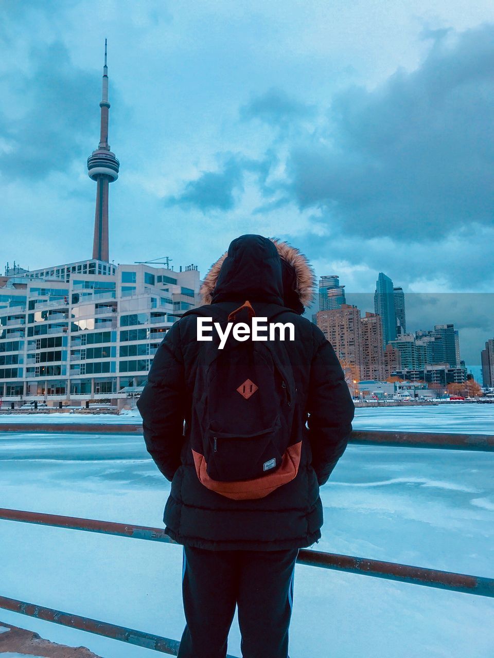 REAR VIEW OF MAN STANDING BY BUILDINGS AGAINST SKY IN CITY