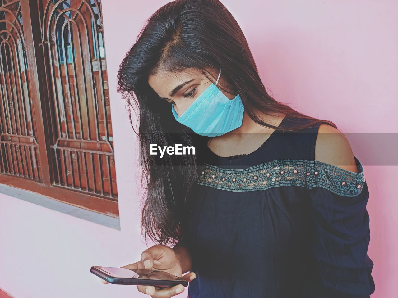 Young woman standing against wall checking her phone wearing surgical mask 