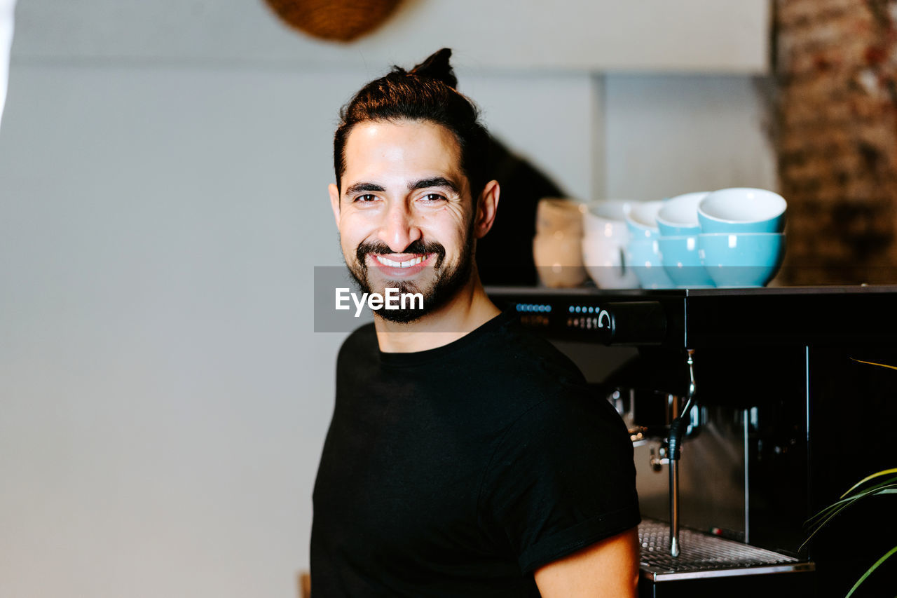 Smiling male barista using portafilter and preparing coffee in modern coffeemaker while standing at counter in cafe and looking at camera