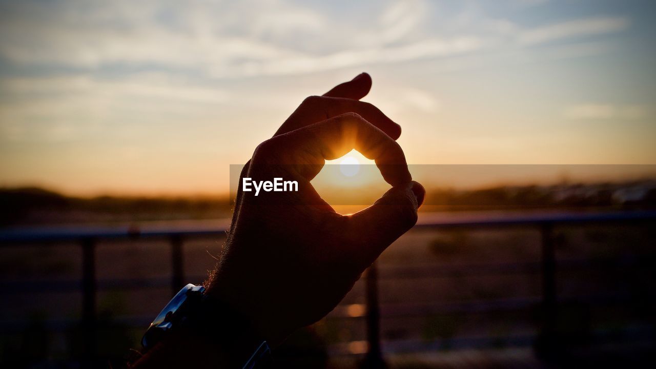 Cropped hand showing ok sign against sky during sunset