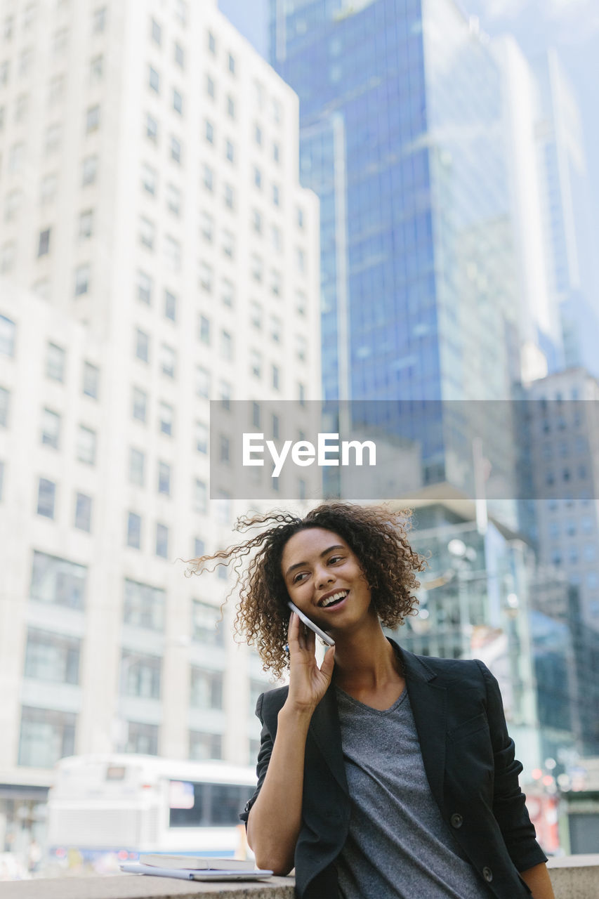 Businesswoman talking on mobile phone while standing in front of office buildings in city