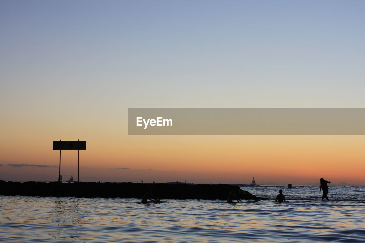 Silhouette people in sea against orange sky during sunset