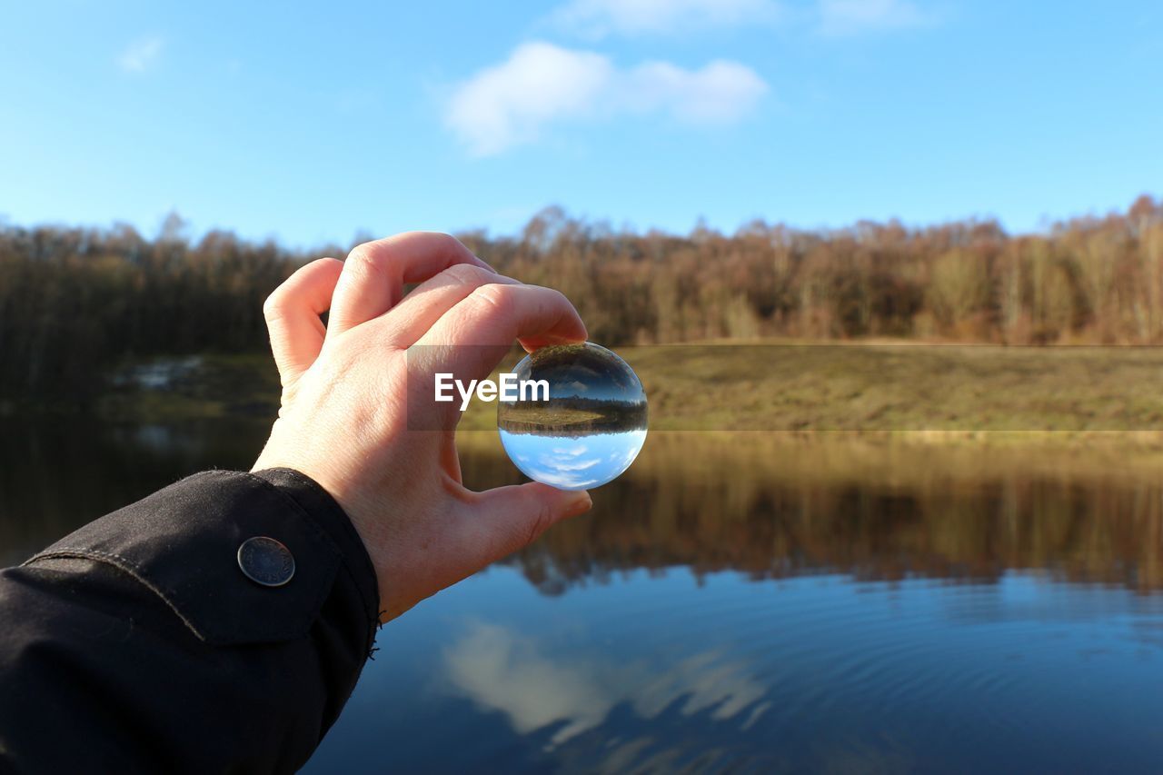 Cropped hand holding crystal ball over lake against trees