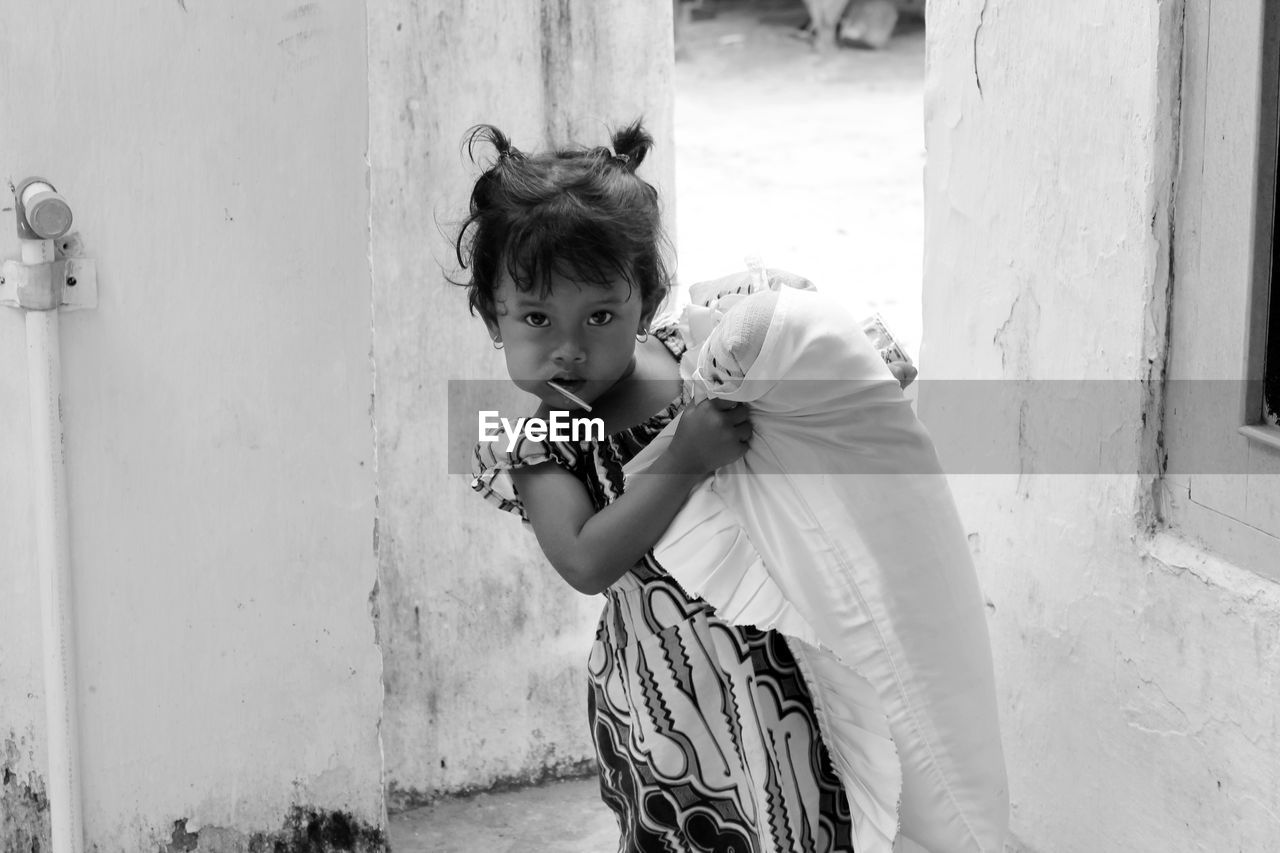 Portrait of girl having lollipop and carrying pillow against built structure