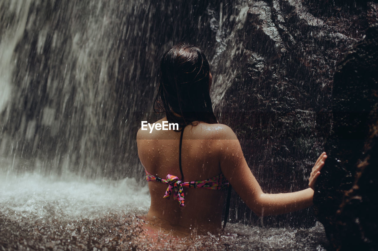Rear view of sensuous young woman standing in river against waterfall