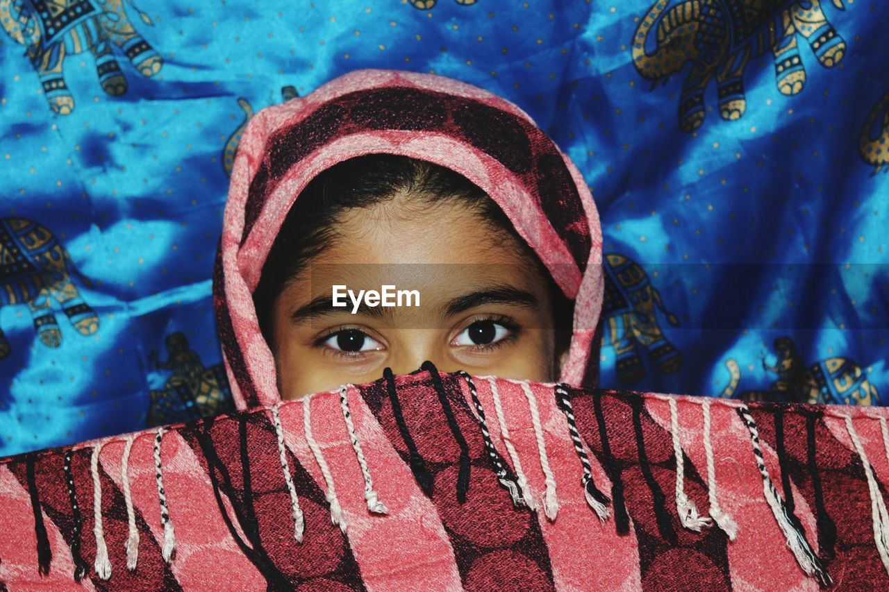 Portrait of girl covering face with scarf against blue fabric