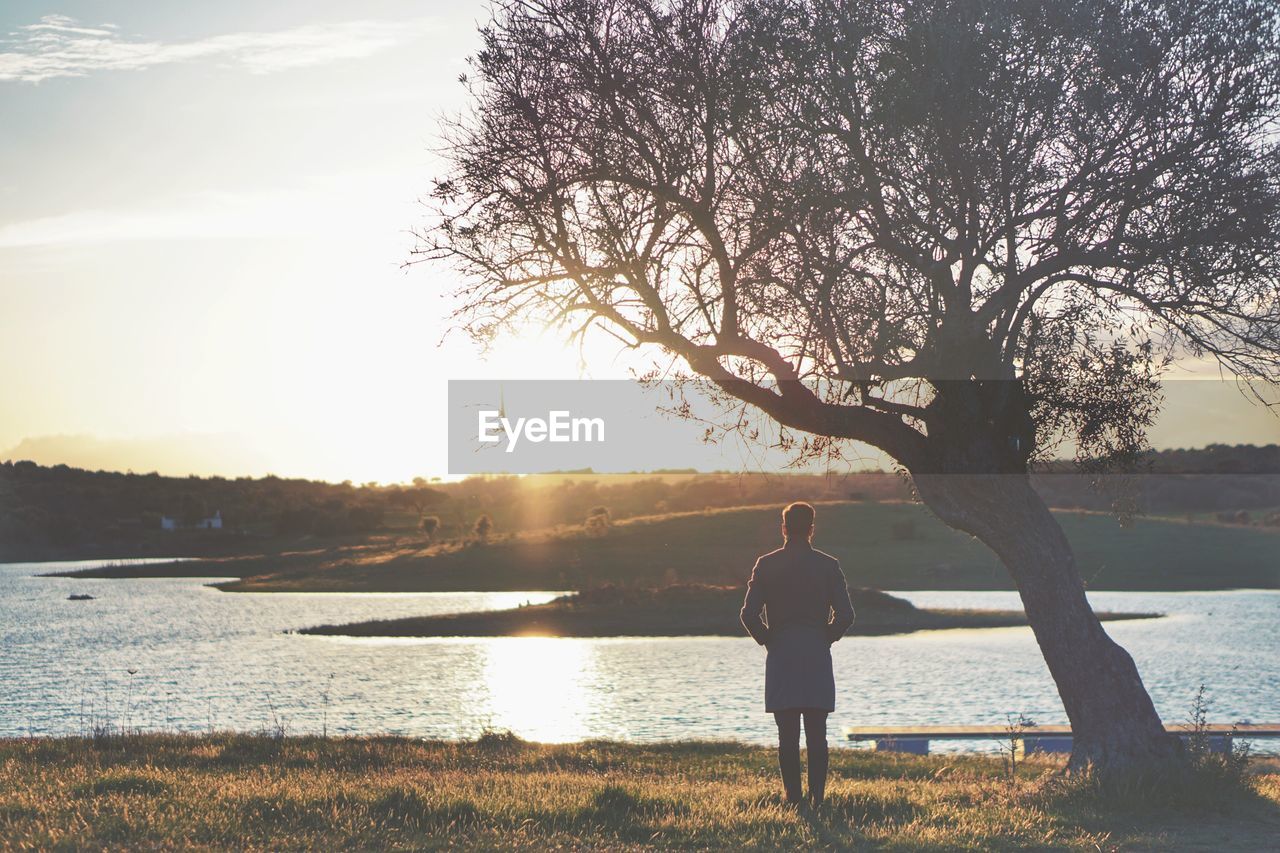 Rear view of man standing by river against sky during sunset