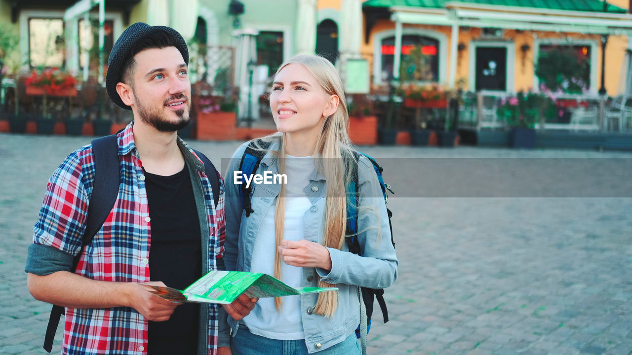 SMILING YOUNG COUPLE STANDING AGAINST CITY IN BACKGROUND