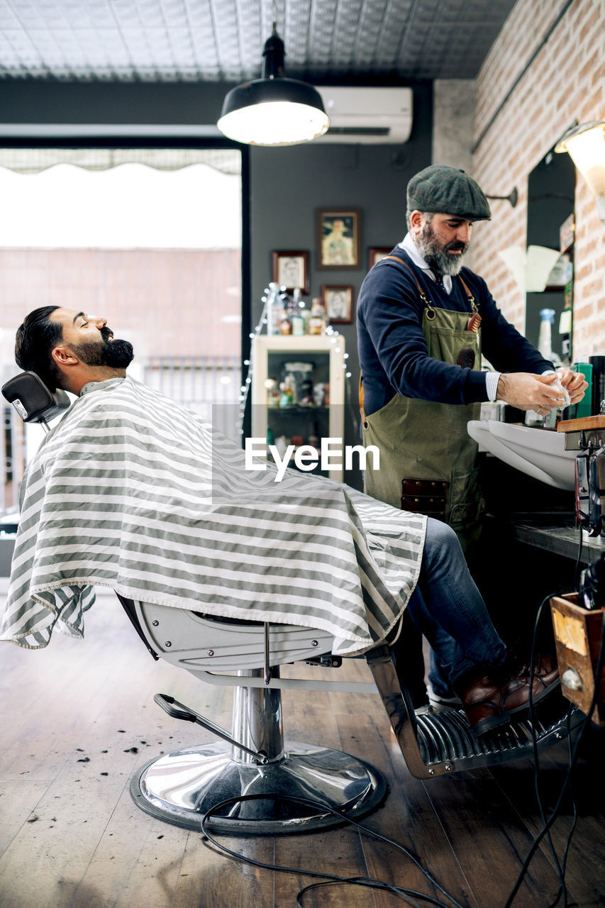 Side view of positive young bearded hispanic male client sitting on chair near middle aged barber preparing for haircut in traditional barbershop