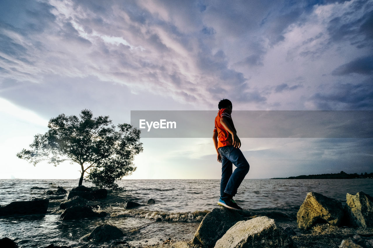 Boy standing on rock by sea against sky