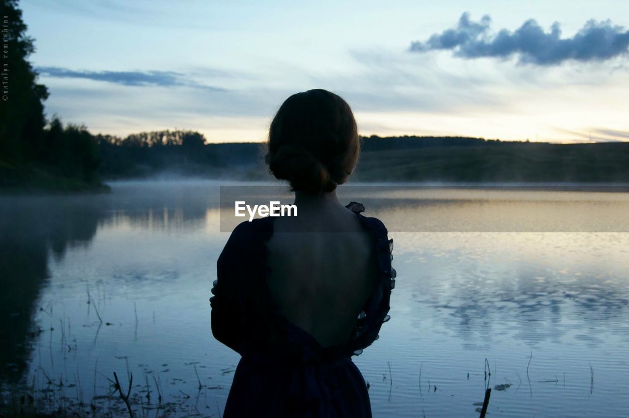 Rear view of woman standing at lakeshore against sky during sunset