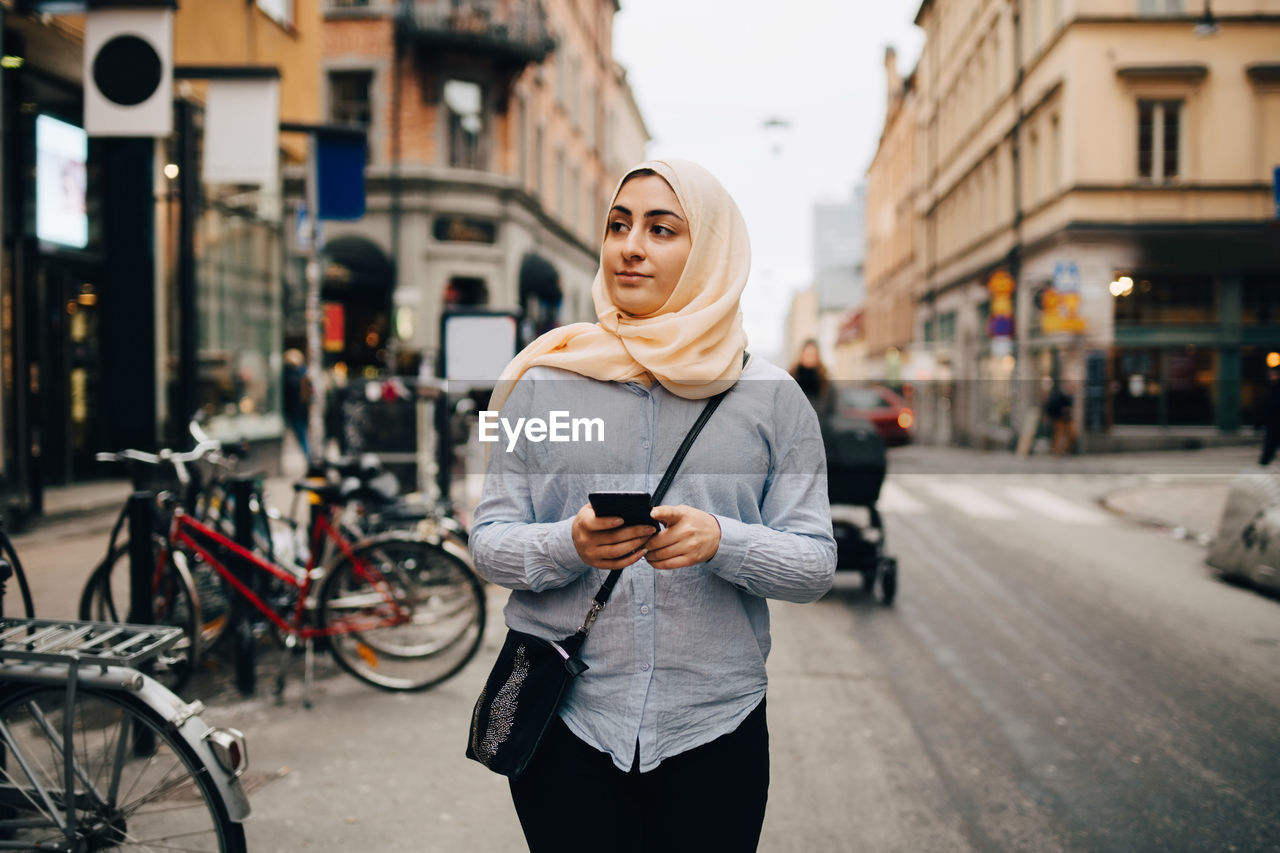 Young muslim woman looking away holding smart phone while walking on street in city