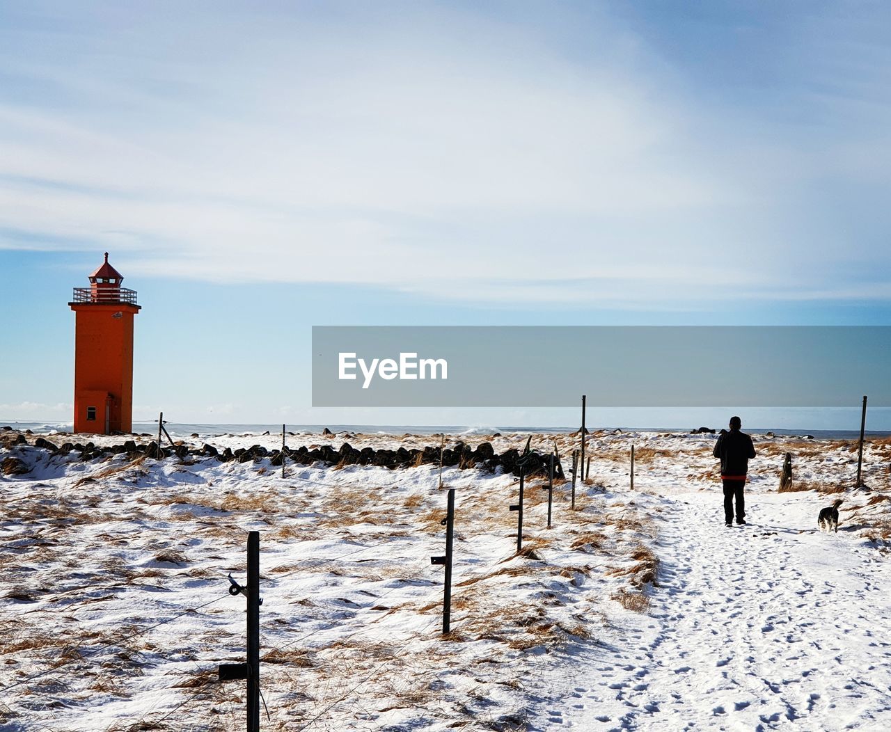 Rear view of man walking on snow covered road leading towards lighthouse against sky during winter