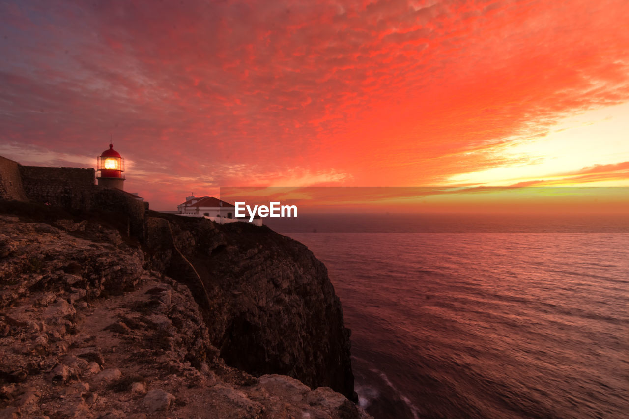 Coastline landscape at sunset with reddish sky and a lighthouse lit on the cape of san vicente, portugal