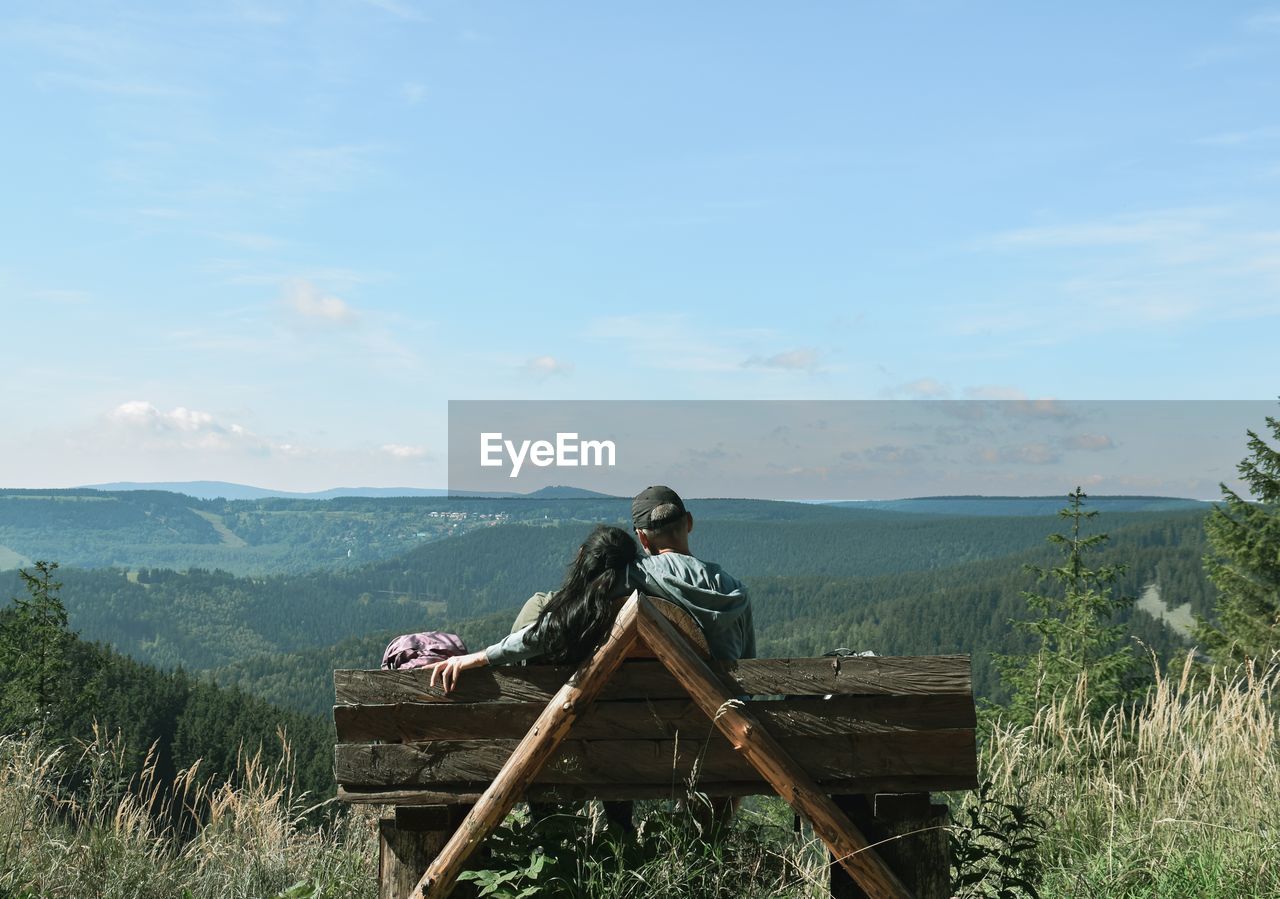 Rear view of couple sitting on wooden bench against sky