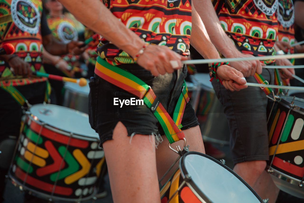 Woman in brightly patterned clothes and black shorts playing steel drums during a street festival.