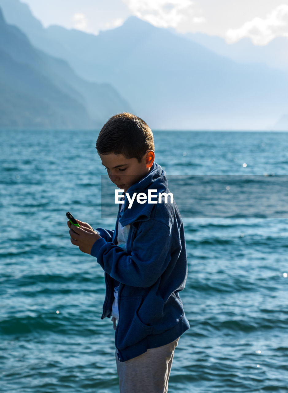 Side view of boy holding mobile phone against sea
