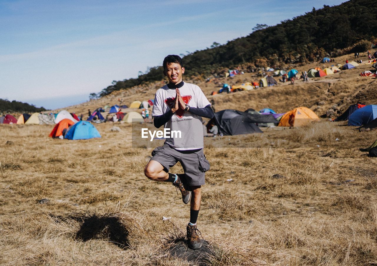 Portrait of young man exercising on grassy field against sky during sunny day at campsite