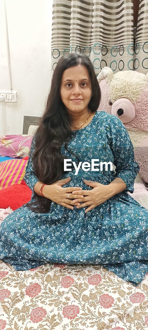 one person, women, sitting, looking at camera, portrait, adult, smiling, front view, indoors, lifestyles, furniture, happiness, long hair, hairstyle, pattern, clothing, young adult, home interior, relaxation, leisure activity, three quarter length, casual clothing, textile, emotion, dress, female, brown hair, full length, black hair, domestic room, art, domestic life, cross-legged