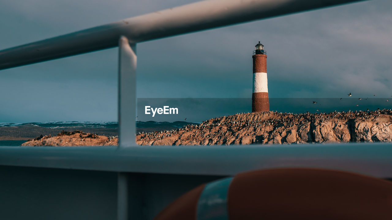 Lighthouse on rock by sea against sky seen through boat railing