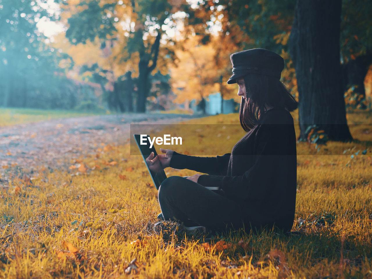 Side view of young woman using laptop while sitting at public park during autumn