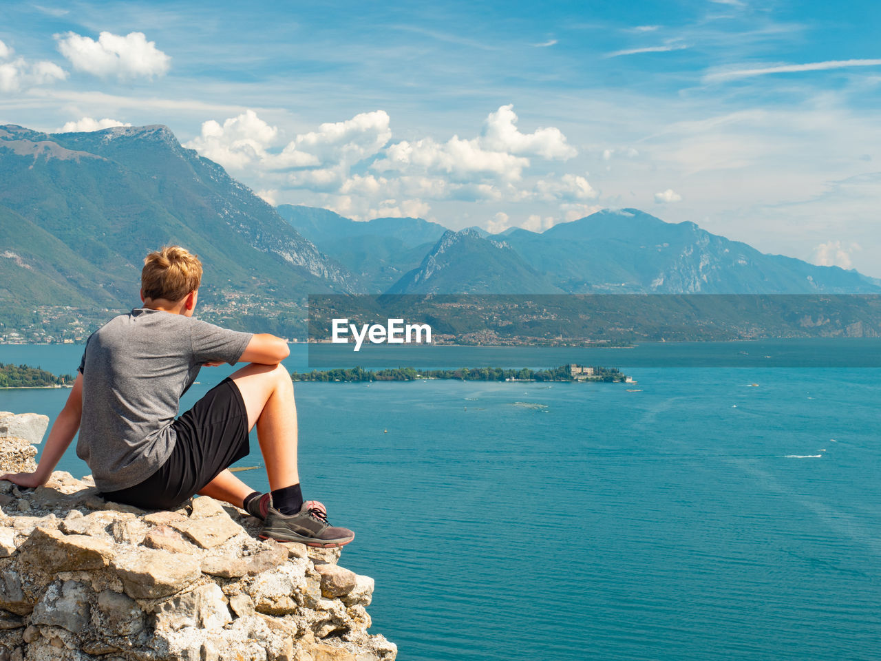 Sports body boy resting on rough stony wall above blue water of garda lake, manerba de rocca, italy