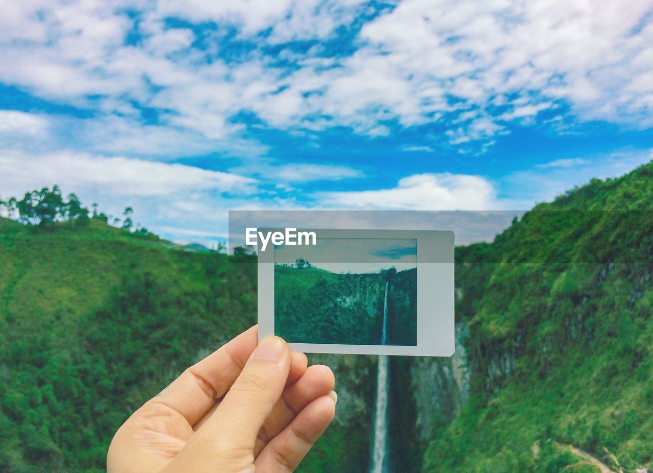 Cropped hand of person holding instant print transfer against waterfall and mountain