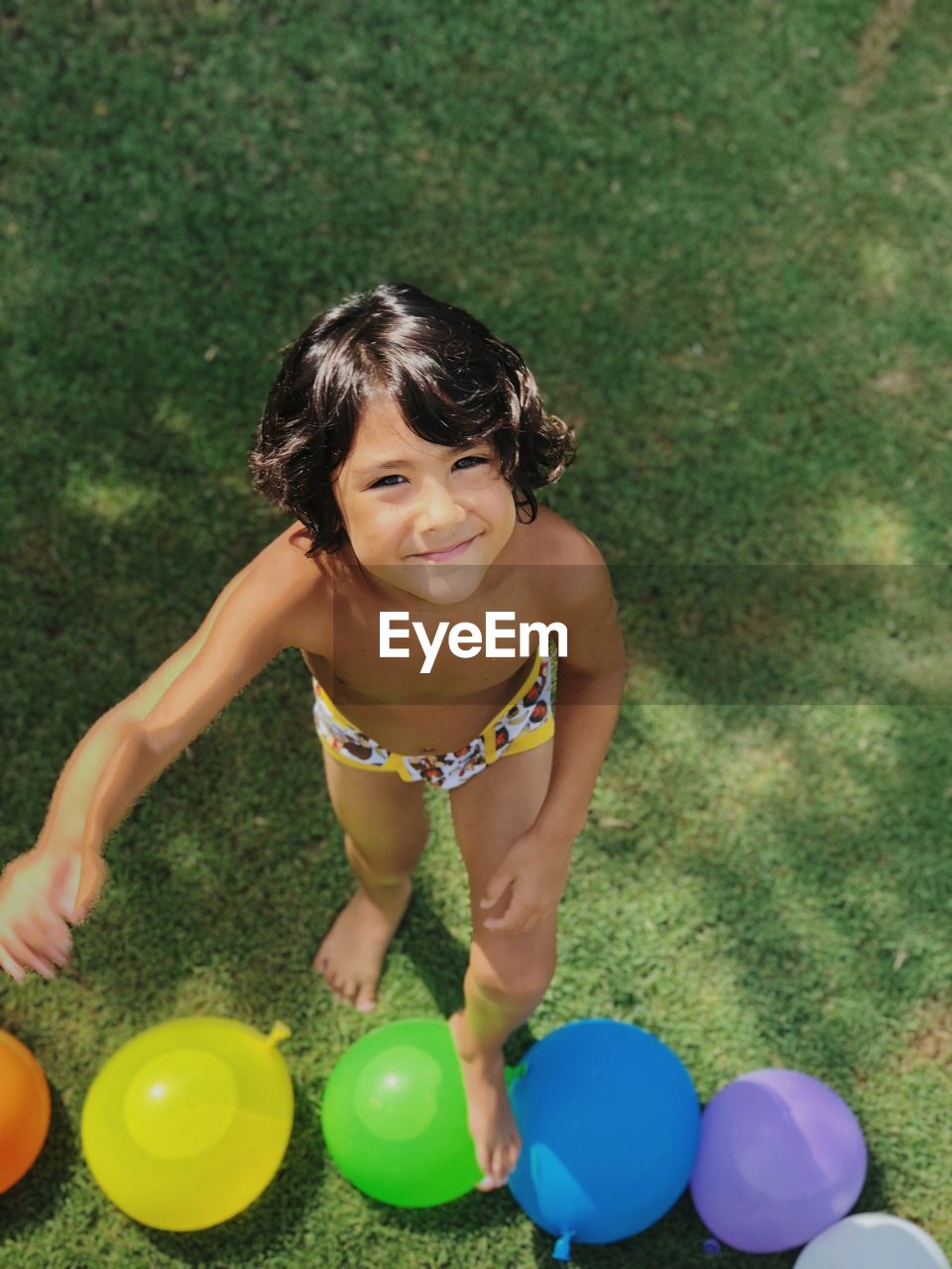 High angle portrait of smiling boy standing by water bomb on grass at back yard