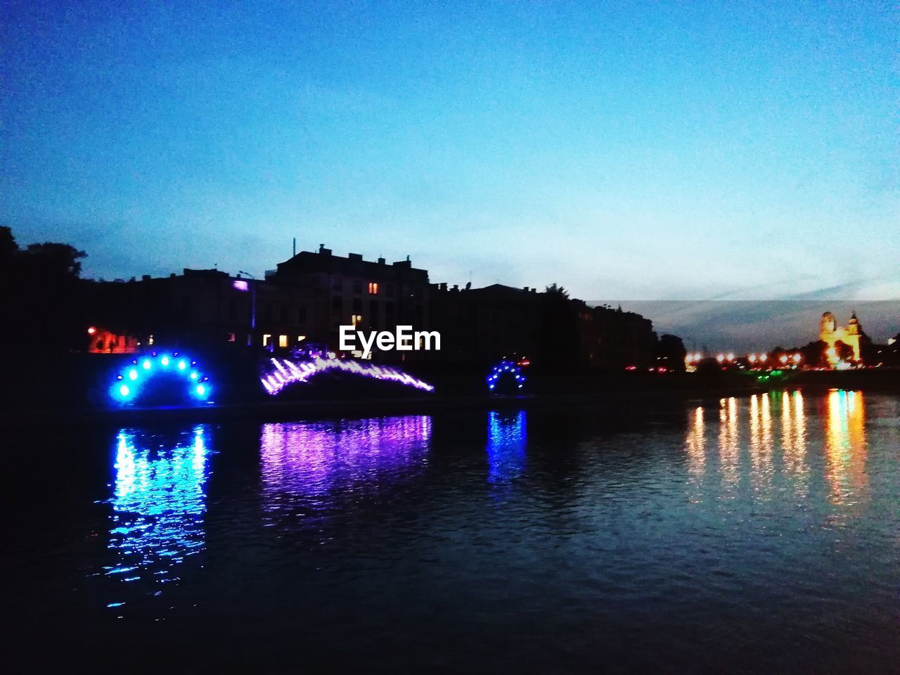 ILLUMINATED BUILDINGS BY RIVER AGAINST SKY AT DUSK