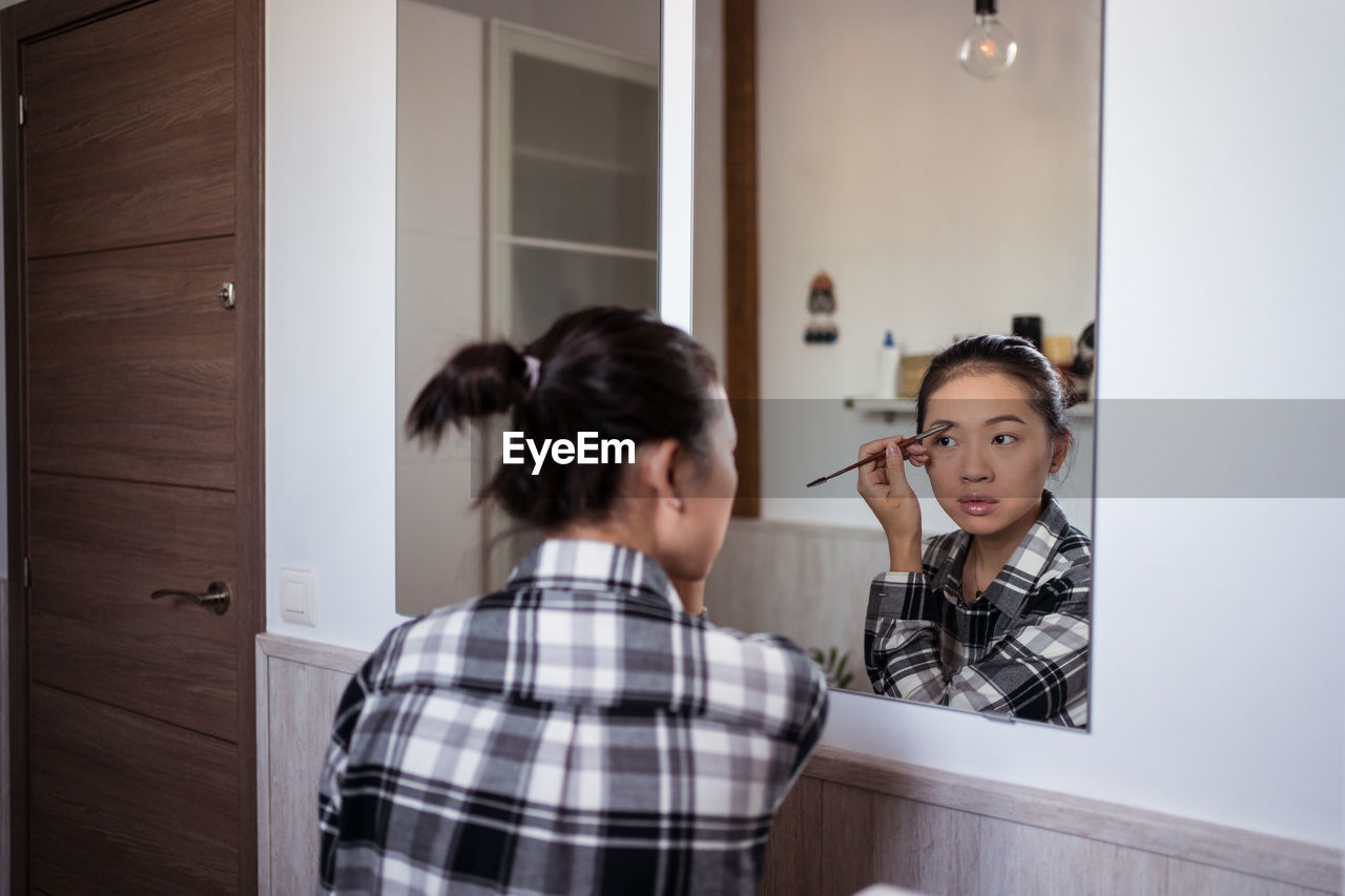 Focused charming ethnic female applying eyebrow pencil while doing makeup and looking in mirror