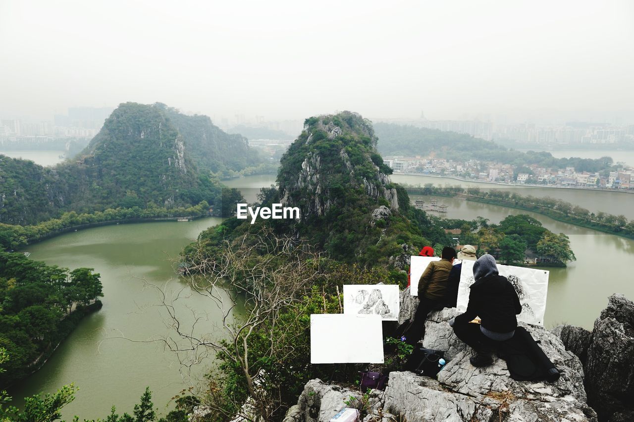Rear view of people painting on canvas against river and mountains during foggy weather