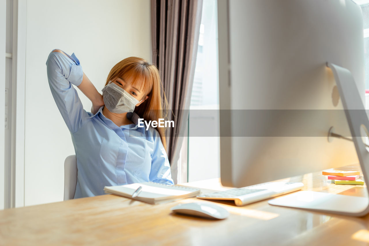 Young woman wearing mask stretching hand while working at home