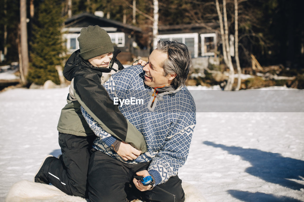 Happy boy in warm clothing embracing father ice fishing during sunny day