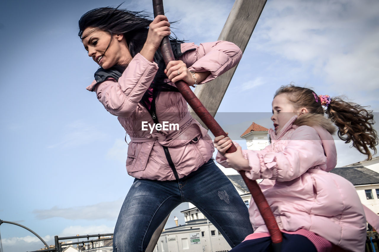 Mother pushing daughter sitting on swing in playground against sky