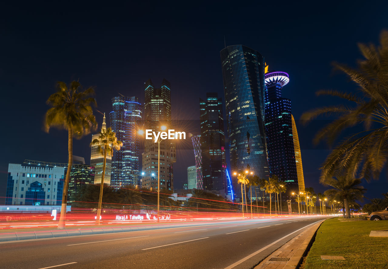 LIGHT TRAILS ON STREET BY ILLUMINATED BUILDINGS AGAINST SKY AT NIGHT