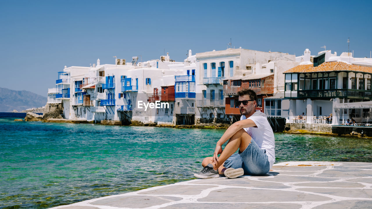 SIDE VIEW OF MAN SITTING AT SEA AGAINST SKY