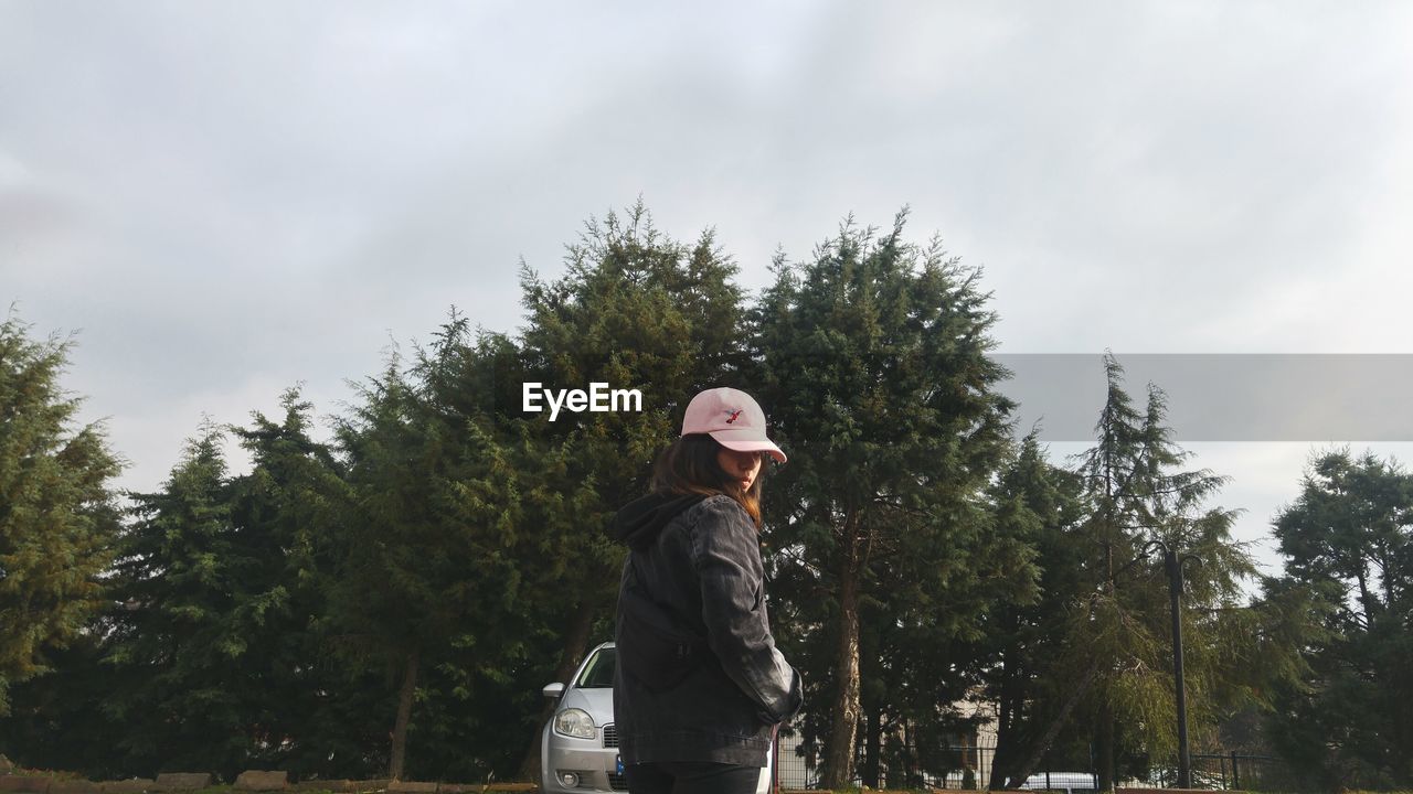 LOW ANGLE VIEW OF WOMAN STANDING BY TREES AGAINST SKY