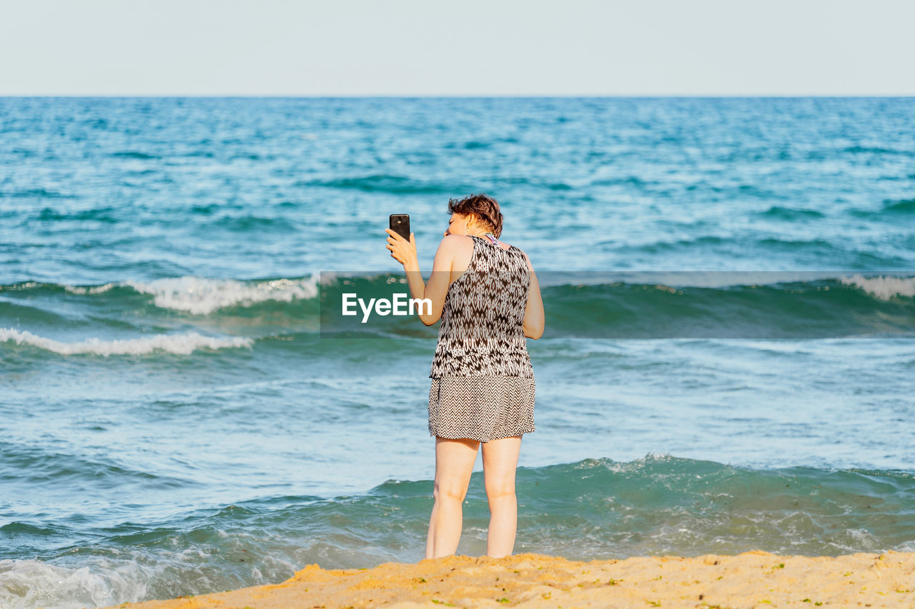 REAR VIEW OF WOMAN STANDING ON BEACH AGAINST SEA