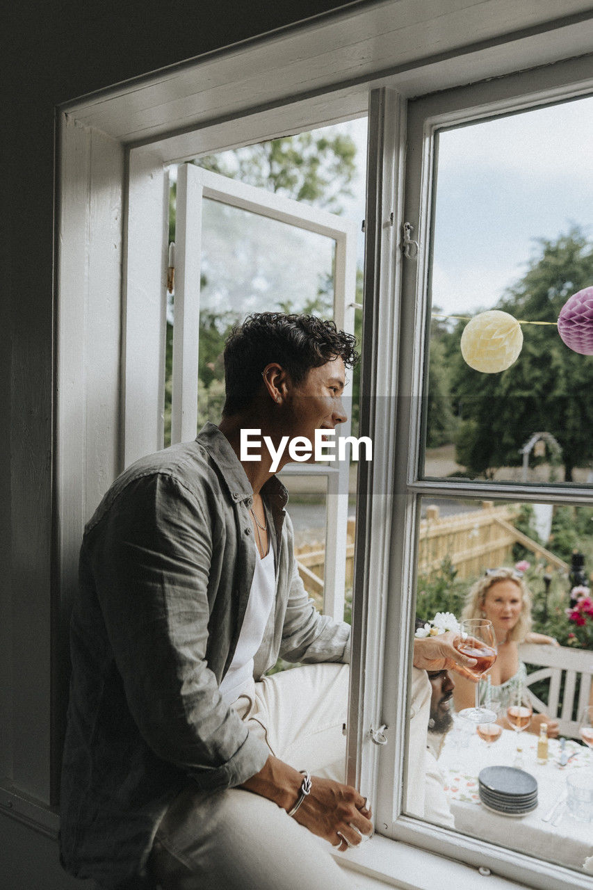 Smiling young man sitting on window sill while looking at friends celebrating party at cafe